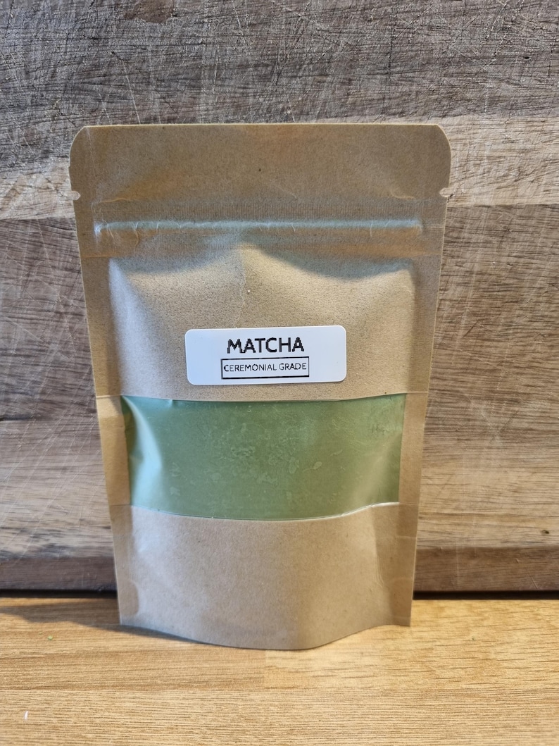 Pu&ograve; includere: Una busta di carta kraft con una finestra trasparente che mostra polvere di matcha verde. Un'etichetta bianca sulla parte anteriore recita "MATCHA CEREMONIAL GRADE". La busta &egrave; sigillata.
