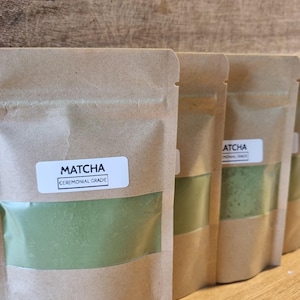 May include: Several brown paper pouches of matcha powder are arranged in a row. Each pouch has a clear window showing the vibrant green powder. The label on the front of the pouch reads "Matcha Ceremonial Grade."