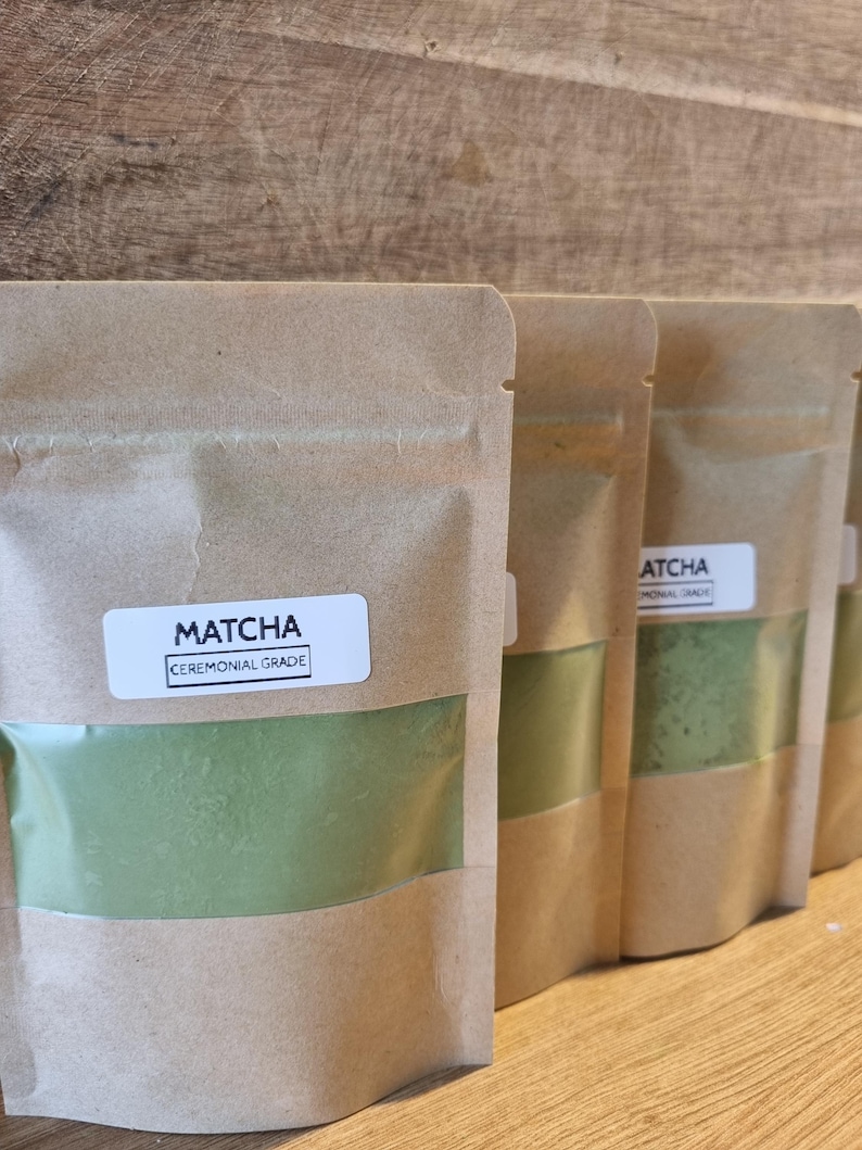 Pu&ograve; includere: Diverse bustine di carta marrone contenenti polvere di matcha sono allineate. Ogni bustina ha una finestra trasparente che mostra la polvere verde brillante. L'etichetta frontale recita "Matcha Ceremonial Grade".