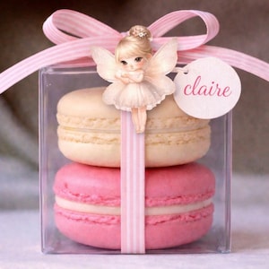 May include: A clear box tied with a pink ribbon, containing two macarons. One macaron is pink, the other is cream-coloured. A fairy figurine and a tag with the name "claire" are attached to the box.
