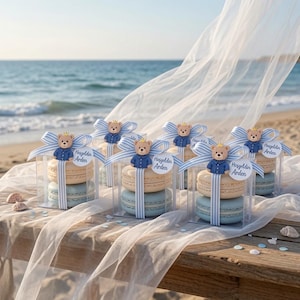 May include: Clear square boxes containing two stacked macarons, one beige and one light blue. Each box is wrapped with a blue and white striped ribbon, topped with a small teddy bear wearing a blue shirt and a gold crown, and a tag reading Hosgeldin Arden.