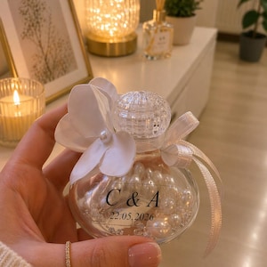 May include: A clear, round glass favor filled with white pearls, adorned with a white flower and a ribbon. The bottle is labeled with the initials "C & A" and the date "22.05.2026".