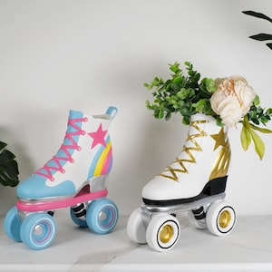 May include: Two decorative roller skate planters. The left skate is white with light blue accents, pink laces, a pink star, and a rainbow graphic. The right skate is white with gold laces, a gold star, and gold heel detail, containing a floral arrangement.