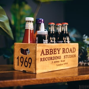 May include: A rectangular wooden crate with cut-out handles, featuring the text ABBEY ROAD RECORDING STUDIOS 3 Abbey Road London NW8 and 1969 printed in dark brown. The crate holds several glass bottles and a squeeze bottle with a white cap.