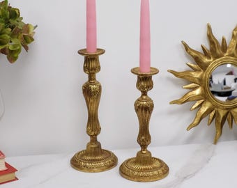 May include: Two gold-toned metal candle holders with ornate, textured detailing on the stems and circular bases. Each holder features a tall, slender, solid pink taper candle inserted into the top cup, creating a pair of decorative lighting pieces.