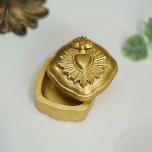 May include: A small, open, gold-coloured trinket box. The lid features a heart-shaped design with sunburst rays. The box is square with rounded edges. The box is likely made of metal or a similar material.