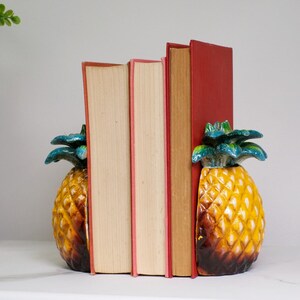May include: A pair of pineapple-shaped bookends with yellow and brown textured bodies and green leafy tops, supporting three books with red spines and cream-coloured pages, arranged vertically between the two decorative pineapple halves.