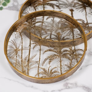 May include: Two round serving trays with metallic gold-toned rims and a distressed finish. The clear glass bases feature a detailed gold-coloured print of palm trees and foliage, creating a layered tropical design across both circular surfaces.