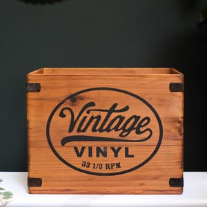 May include: A rectangular wooden storage crate with a medium brown finish and dark metal corner accents. The front features a black oval graphic containing the text Vintage VINYL 33 1/3 RPM in a combination of script and block lettering.