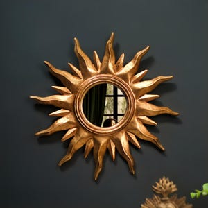 May include: A gold sunburst mirror with a circular reflective centre. The mirror has a textured, metallic finish and is mounted on a dark wall. The sun's rays are stylized and pointed.
