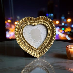 May include: A heart-shaped, gold picture frame with a scalloped edge. The frame contains a silhouette image against a backdrop of handwritten script. The frame is displayed on a reflective surface, with a blurred city skyline in the background.