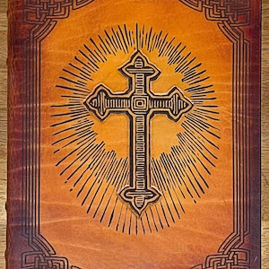 May include: A brown leather-bound journal with a cross design embossed on the front cover. The cross is surrounded by a sunburst pattern and the edges of the cover are decorated with a Celtic knot design.