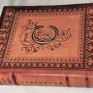 May include: A brown leather-bound Holy Bible with a gold-colored initial "C" and ornate floral designs on the cover. The spine reads "HOLY BIBLE CARPENTER".