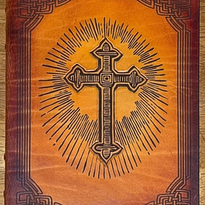 May include: A brown leather-bound journal with a cross design embossed on the front cover. The cross is surrounded by a sunburst pattern and the edges of the cover are decorated with a Celtic knot design.