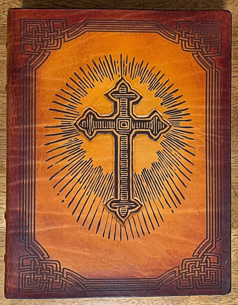 May include: A brown leather-bound journal with a cross design embossed on the front cover. The cross is surrounded by a sunburst pattern and the edges of the cover are decorated with a Celtic knot design.
