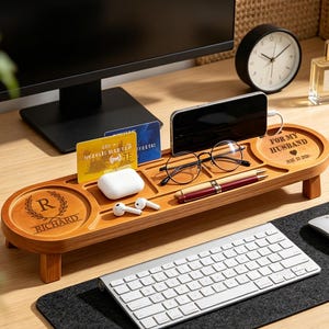Op de afbeelding: Een houten bureau-organizer met meerdere compartimenten. Het bevat een smartphone, bril, pennen, creditcards en andere kleine items. De organizer heeft de tekst "FOR MY HUSBAND" en "RICHARD" gegraveerd. Een toetsenbord, muis en klok zijn ook zichtbaar.
