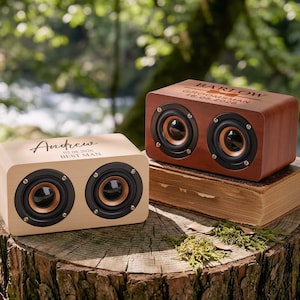 Op de afbeelding: Twee houten luidsprekers met zwarte en gouden luidsprekerconussen. Eén luidspreker is van licht hout met de naam "Andrew" en "Best Man" gegraveerd. De andere is van donker hout met de naam "Barlow" en "Groomsman" gegraveerd.