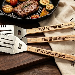 May include: Three stainless steel grilling spatulas with wooden handles. The spatulas have the phrases "BEST FLIPPIN'DAD Love, Kevin", "The GrillFather Love, Ashley", and "BEST FLIPPIN'GRANPA Love, Grace" engraved on the handles. A cast iron pan with grilled food is in the background.