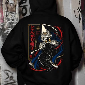Könnte beinhalten: Ein schwarzer Kapuzenpullover mit einer Grafik auf der Rückseite. Das Design zeigt eine Figur mit spitzem Hut und einem zentralen Auge, die einen blauen Umhang und dunkle Kleidung trägt. Japanischer Text und Atelier of Witch Hat sind abgebildet.