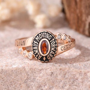 May include: A rose gold-toned ring featuring a central oval-cut reddish-brown gemstone surrounded by a black border with the text ALWAYS MOVING FORWARD. The band is engraved with 2025 and CHRISTOPHER, accented with small clear stones on the sides.