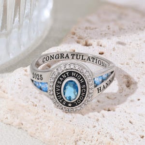 May include: Silver-toned class ring with a central oval blue gemstone, surrounded by a halo of small clear stones. The ring features the inscription "CONGRATULATIONS" and "STUYVESANT HIGH SCHOOL". Additional text includes "2025" and a name.