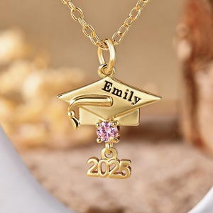 Op de afbeelding: Goudkleurige afstudeerpet hanger ketting met de naam "Emily" en het jaar "2025" gegraveerd. De hanger heeft een roze edelsteen en hangt aan een delicate ketting.