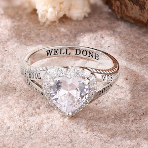 May include: A silver ring featuring a heart-shaped, clear gemstone encircled by smaller clear stones. The ring is inscribed with "WELL DONE", "Olivia", "WESTERN", and "2025". The ring rests on a sandy surface.