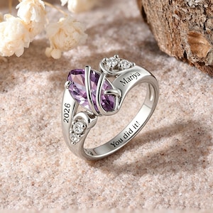 May include: A silver ring with a large, oval-cut purple gemstone. The ring features the engraved text "2026", "Mariya", and "You did it!". Small clear stones accent the design.