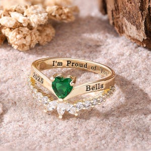 May include: Gold-toned ring with a heart-shaped green gemstone and the inscription "I'm Proud of You!" and "Bella". The ring features a floral design with clear gemstones and the year "2025".