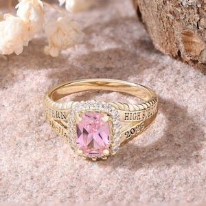 Könnte beinhalten: Ein goldfarbener Ring mit einem rechteckigen, rosafarbenen Edelstein in der Mitte, umgeben von kleineren, klaren Steinen. Auf dem Ring steht "Western Isabella High School 2026".