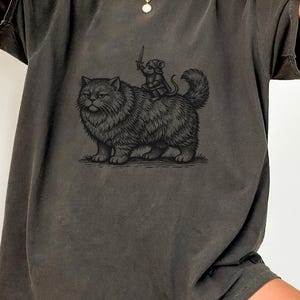 May include: Dark grey t-shirt featuring a black illustration of a fluffy cat with a mouse riding on its back, holding a sword. The shirt has short sleeves and a relaxed fit, suitable for casual wear.