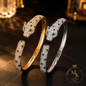 Könnte beinhalten: Zwei offene Armreifen in Form von Pantherköpfen, einer in Gold- und einer in Silberoptik. Beide sind mit klaren Pavé-Steinen, schwarzen Flecken und grünen Augen besetzt. Ein Logo mit dem Text MK Création Shop befindet sich in der Ecke.