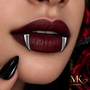 Pode incluir: Um par de piercings labiais prateados, curvos e em forma de presa, posicionados no lábio inferior. A joia apresenta um acabamento metálico liso e polido com um design pontiagudo. O texto MK création é visível no canto inferior direito.