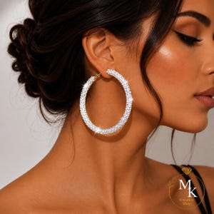 May include: A large circular hoop earring featuring a pavé setting of clear, reflective crystals. The jewelry piece has a metallic base and a continuous, uniform surface covered in small, sparkling stones that catch the light.