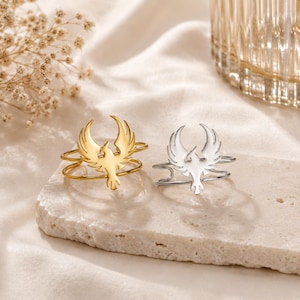 May include: Two rings with a phoenix design. One ring is gold-colored, and the other is silver-colored. The phoenix is the focal point of each ring, with its wings spread wide. The rings are displayed on a light-colored, textured surface.