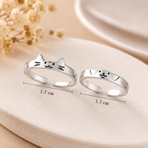 Può includere: Due anelli regolabili color argento, uno con un design a forma di gatto e l'altro con un design a forma di cane. Ogni anello misura 1,7 cm di lunghezza. Gli anelli sono esposti su una superficie chiara.
