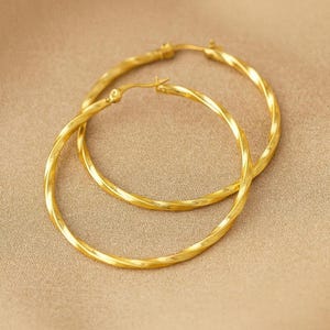 Könnte beinhalten: Ein Paar goldfarbene Creolen mit einem gedrehten Seildesign. Die Ohrringe werden auf einer neutralen Oberfläche präsentiert, die ihre helle, reflektierende Oberfläche zeigt. Die Creolen sind ein klassisches Schmuckstück.