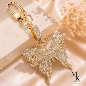 Peut inclure: Un porte-clés en métal doré avec un pendentif papillon incrusté de strass transparents. Le papillon est fixé à un fermoir mousqueton doré par un court connecteur rectangulaire également recouvert de strass. Le logo MK Création Shop est visible.