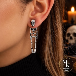 Peut inclure: Une boucle d'oreille argentée composée d'un clou en forme de crâne relié à un corps de squelette pendant. Le design présente des détails oxydés pour accentuer la structure osseuse, incluant la cage thoracique, le bassin et les os des jambes.