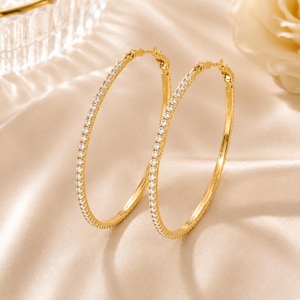 Peut inclure: Une paire de créoles dorées. Un côté de chaque boucle d'oreille est incrusté de petites pierres précieuses transparentes. Les boucles d'oreilles sont présentées sur une surface beige satinée. Les boucles d'oreilles ont un design classique et élégant.