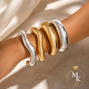 Peut inclure: Un ensemble de quatre bracelets en métal épais portés au bras. La collection comprend deux bracelets argentés au design lisse, ondulé ou incurvé et deux bracelets dorés avec une finition épaisse, arrondie et polie.