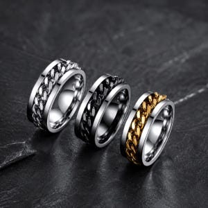 Peut inclure: Trois bagues en acier inoxydable avec un motif de chaîne. Une bague est argentée, une est noire et la troisième a une chaîne dorée. Les bagues sont présentées sur une surface sombre.