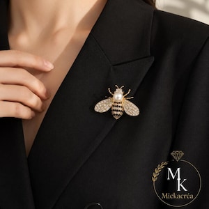 Peut inclure: Une broche abeille dorée avec une tête en perle blanche, des rayures de corps incrustées de cristaux noirs et des ailes pavées de cristaux clairs. L'épingle en forme d'insecte est fixée sur un blazer noir. Le logo Mickacréa est visible en bas.