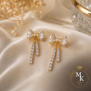 Peut inclure: Une paire de boucles d'oreilles dorées en forme de nœud avec des cristaux transparents à facettes. Chaque boucle présente un nœud central avec deux brins pendants incrustés de cristaux. Le logo MK Creation Shop est visible en bas à droite.