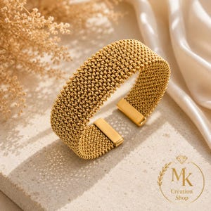Peut inclure: Un bracelet manchette doré présentant un design en maille tissée texturée avec des embouts métalliques lisses et polis. Le bracelet possède une structure ouverte, mettant en valeur un motif complexe et flexible qui entoure le poignet.