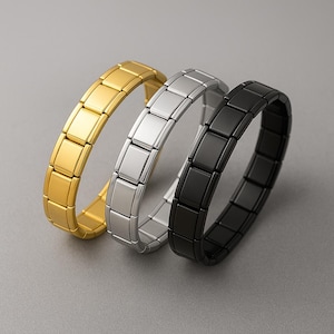 May include: Three bracelets in gold, silver, and black. Each bracelet is composed of rectangular links, creating a flexible, curved shape. The bracelets are displayed on a gray surface, showcasing their metallic sheen.