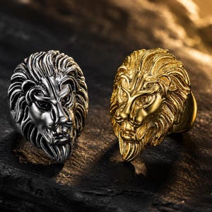 Puede incluir: Dos anillos con diseños de cabeza de león. Uno es plateado, el otro dorado. Las cabezas de león tienen melenas y rasgos faciales detallados. Los anillos se muestran sobre una superficie oscura y texturizada.