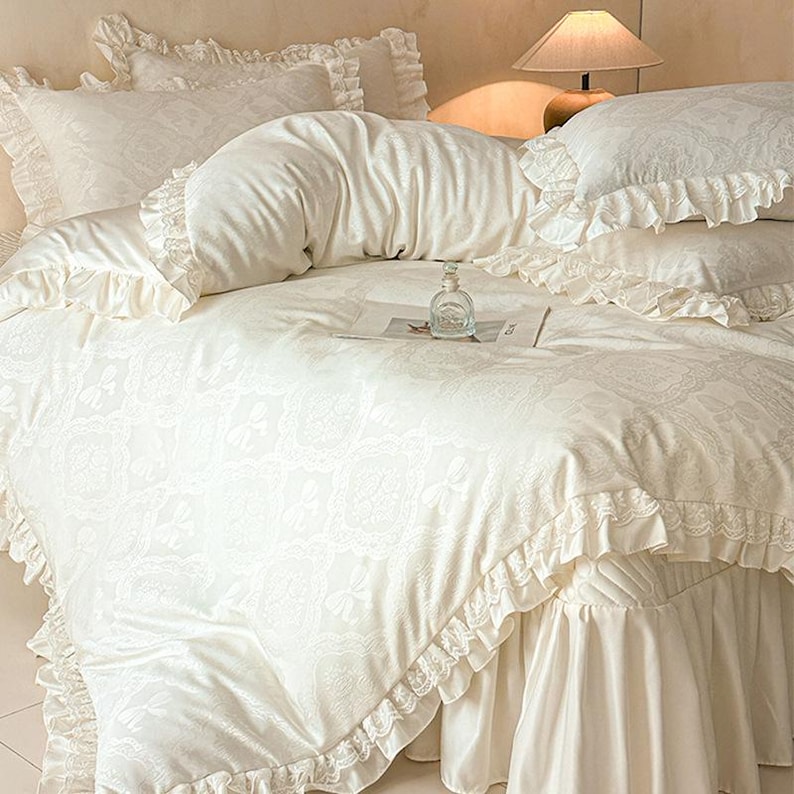 K&ouml;nnte beinhalten: Ein cremefarbenes Bettw&auml;sche-Set bestehend aus einem Bettbezug und passenden Kissenbez&uuml;gen mit einem dezenten Jacquard-Muster aus Schleifen und spitzenartigen R&auml;ndern. Die Kanten sind mit einem R&uuml;schenbesatz aus Stoff versehen.
