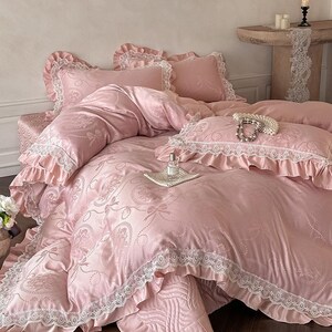 Puede incluir: Un juego de cama rosa claro con un patr&oacute;n jacquard de lazos y motivos florales. La funda n&oacute;rdica y las fundas de almohada tienen volantes de encaje blanco. Un frasco de vidrio peque&ntilde;o descansa sobre una bandeja cuadrada blanca con un collar de perlas.