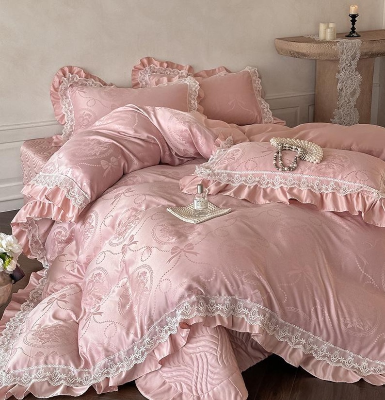 Puede incluir: Un juego de cama rosa claro con un patr&oacute;n jacquard de lazos y motivos florales. La funda n&oacute;rdica y las fundas de almohada tienen volantes de encaje blanco. Un frasco de vidrio peque&ntilde;o descansa sobre una bandeja cuadrada blanca con un collar de perlas.